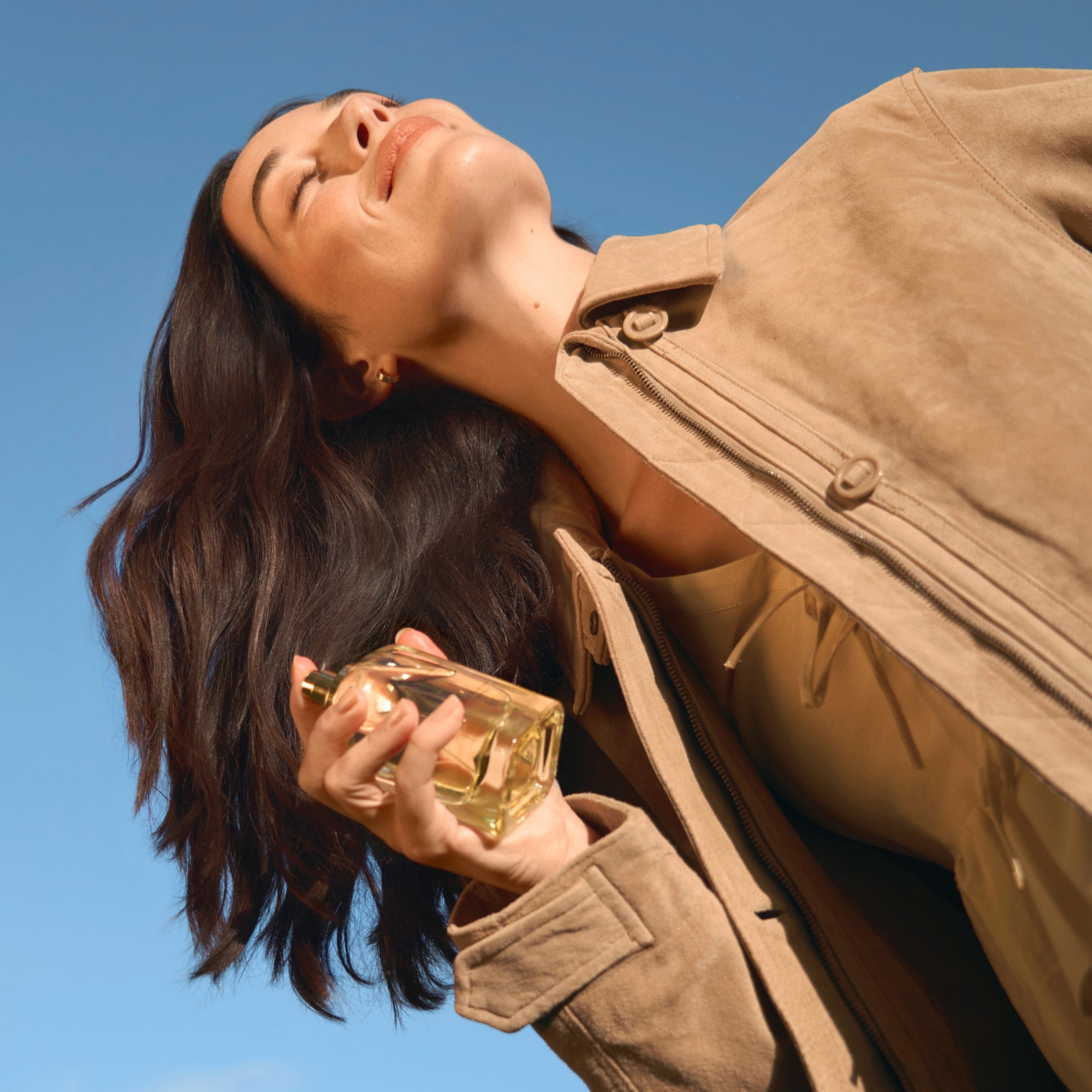 Woman in a beige coat holding a joy of primrose perfume bottle against a clear blue sky.