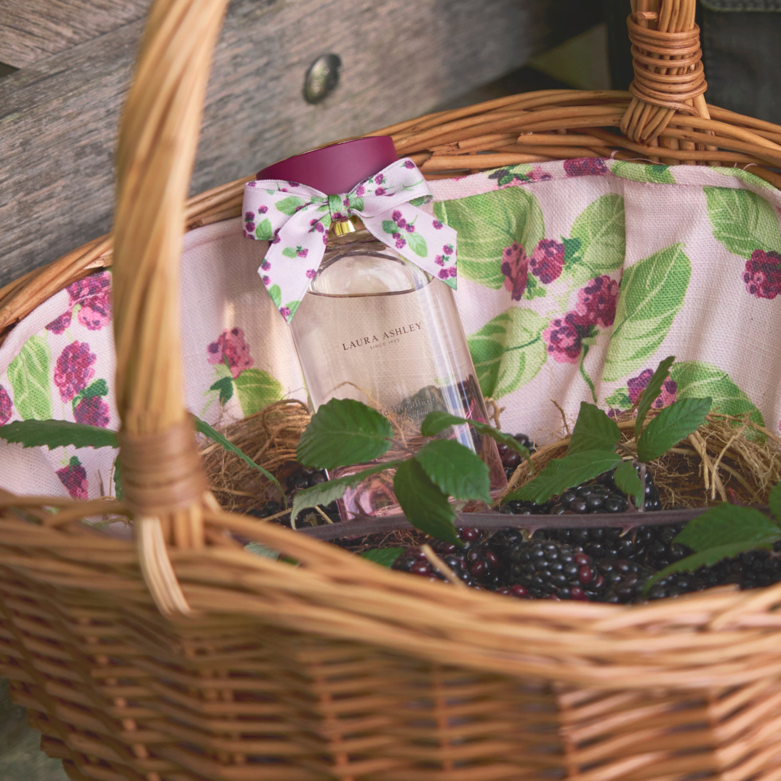 Wicker basket with a floral liner containing a bottle of Laura Ashley perfume and blackberries.